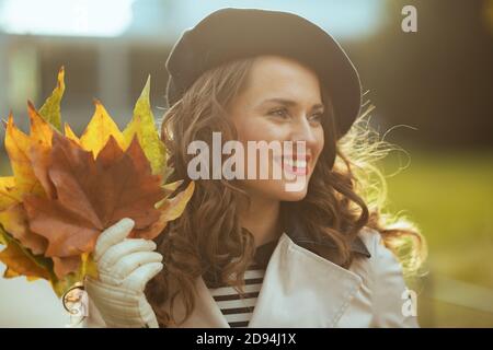 Ciao novembre. Felice donna moderna di 40 anni in trench camice beige e beretto nero con foglie gialle autunnali all'aperto in città in autunno. Foto Stock