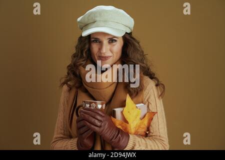 Ciao ottobre. Ritratto di sorridente elegante casalinga di mezza età in sciarpa con foglia di acero giallo autunno, libro, guanti in pelle e coppa contro bronzo b Foto Stock