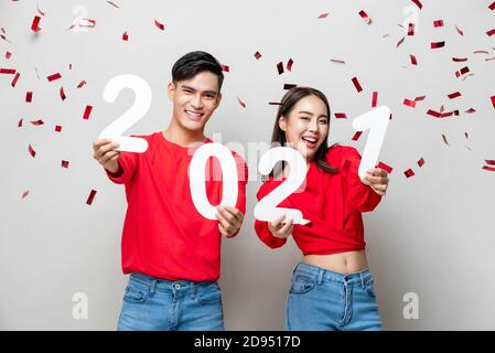 Sorridente felice coppia asiatica in abbigliamento casual rosso 2021 numeri per il concetto di nuovo anno su sfondo grigio chiaro con confetti Foto Stock