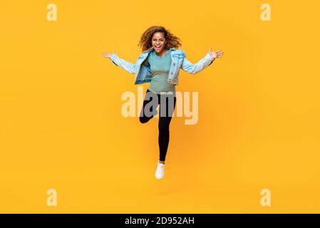 Ritratto a lunghezza intera di giovane energica afroamericana che salta dentro aria media mentre le braccia si allungano in uno studio isolato sfondo giallo Foto Stock