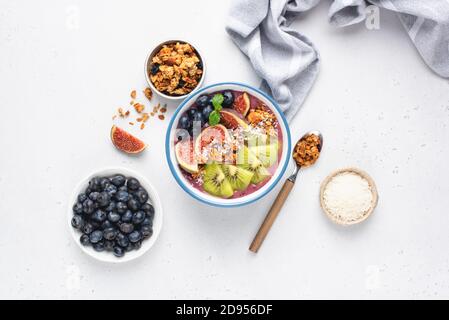 Ciotola di Smoothie di fico di mirtillo con guarnizioni di cocco di kiwi di granola. Piatto posato su sfondo di cemento bianco. Vista dall'alto. Frullato sano e miscelato Foto Stock