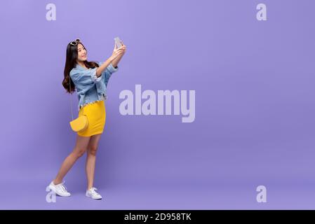 Ritratto a lunghezza intera di sorridente giovane bella donna asiatica prendendo selfie con smartphone in sfondo viola isolato studio con copia spazio Foto Stock