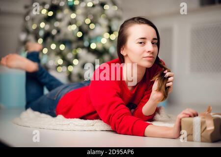 Ritratto di donna bella con dono seduto da albero di Natale a casa Foto Stock
