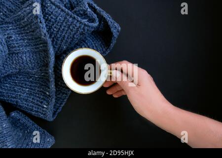 Pullover in maglia blu, una mano di donna tiene una tazza di caffè in porcellana dorata su sfondo nero. Maglieria come concetto di hobby femminile Foto Stock