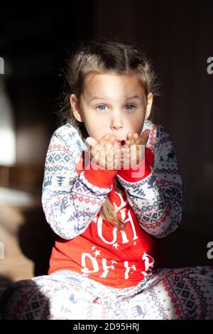 Primo piano ragazza con i pigtail in pigiama di Natale si siede sul pavimento e soffia bacio, mattina di nuovo anno, luce di alba del sole Foto Stock