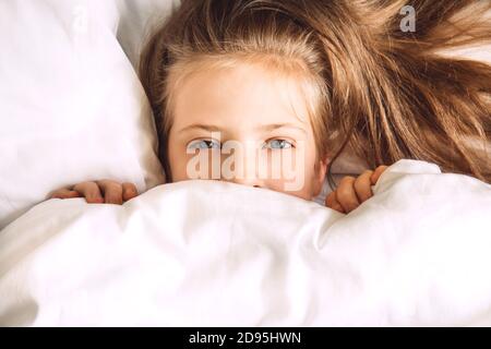 Bambini che si nascondono sotto il piumone nel letto. Routine mattutina. Concedetevi la camera da letto al mattino. Ragazza a letto guardare fuori da sotto le coperture bianche. Messa a fuoco selettiva Foto Stock