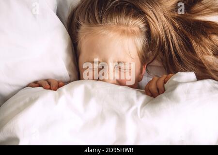 Bambini che si nascondono sotto il piumone nel letto. Routine mattutina. Concedetevi la camera da letto al mattino. La ragazza chiuse gli occhi sotto le coperture fuoco selettivo Foto Stock