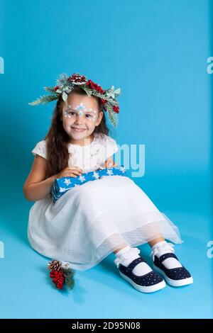 una bella ragazza in un vestito bianco, indossando una corona e sneakers si siede sul pavimento e si apre una scatola di regali, isolato su uno sfondo blu Foto Stock