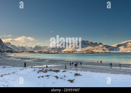 I turisti che visitano la famosa spiaggia di sabbia vicino a Ramberg sulla Lofoten isole in Norvegia in chiaro giorno d'inverno con neve-rivestito montagne e cielo blu Foto Stock