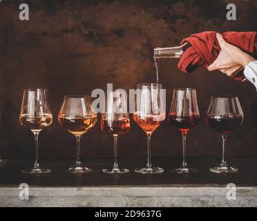 Sfumature di vino Rosa in calici con gambo messi in linea e womans mano versando il vino da bottiglia a bicchiere, arrugginito fondo marrone dietro. Enoteca, enoteca, degustazione Foto Stock