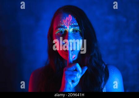 Silenzio e hush concetto. Bella donna con trucco fluorescente tiene il dito vicino alle labbra. Vernice fluorescente in luce al neon. Foto Stock