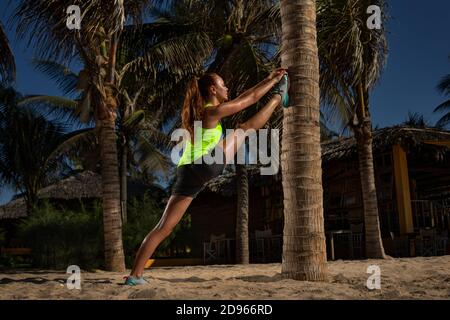 La giovane donna sta facendo delle spaccature vicino ad un albero della palma su una spiaggia Foto Stock