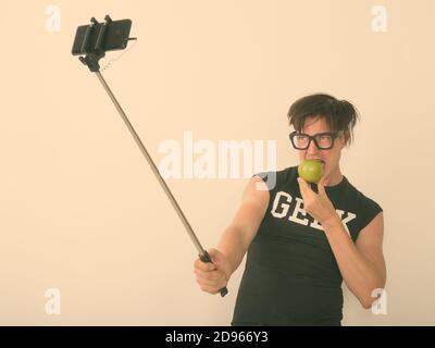 Felice giovane uomo nerd che prende selfie con il telefono su selfie bastone e mangiare mela verde su sfondo bianco Foto Stock