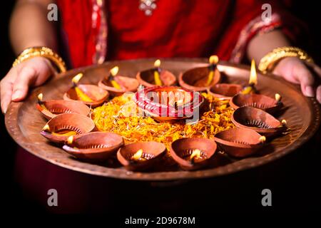 Felice Diwali sfondo. Donna indiana o sposa che indossa tradizionale stoffa rossa e gioielli, tenendo puja thali pieno di luce bruciante diya o lampada ad olio di argilla Foto Stock