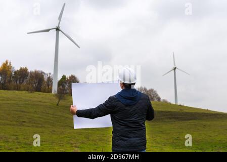 Vista posteriore di un ingegnere maschio indossa il casco bianco che regge un piano a turbina eolica sito autunno nuvoloso cielo verde prato Foto Stock