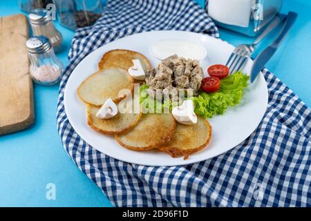 Deruny - frittelle di patate con funghi, champignons, panna acida su un piatto bianco. Pomodori ciliegini, insalata. Il ristorante serve su sfondo blu Foto Stock