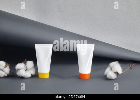 Due tubi cosmetici bianchi con tappi colorati e senza logo accanto ai fiori di cotone su sfondo grigio studio. Primo piano, spazio di copia, simulazione Foto Stock