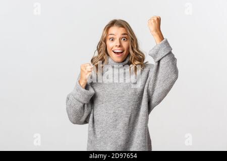 Immagine di ragazza entusiasta di speranza in pullover grigio, guardando sorpreso come vincere e rallegrarsi, alzando la mano in su, in piedi felice contro sfondo bianco Foto Stock