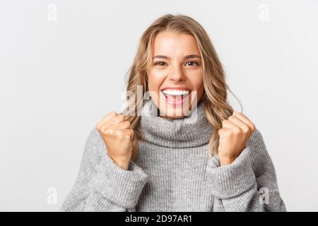 Primo piano di speranzosa ragazza bionda in pullover grigio, pugni clenching e aspetto eccitato, vincendo qualcosa, in piedi su sfondo bianco Foto Stock