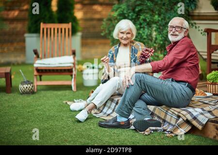 Foto a lunghezza intera di una coppia sorridente che siede insieme sul portico del furgone e mangia le uve. Spazio di copia. Relazione e concetto di famiglia Foto Stock