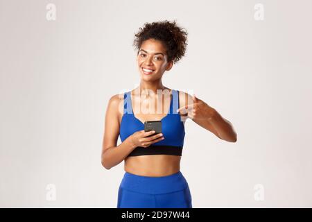 Concetto di sport e tecnologia. Attraente donna afroamericana di idoneità in sport, puntando il dito al telefono mobile, consiglia l'applicazione di allenamento