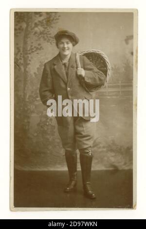 Originale cartolina studio dell'era WW1 di attraente Land Girl nella Women's Land Army (WLA) indossando braghe, giacca e cappello che reggono un cestino di pane, coinvolto nella raccolta del grano, sezione agricola della WLA, datata agosto 1918 sul retro, Regno Unito Foto Stock