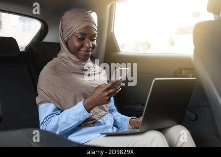 Multitasking. Donna d'affari musulmana nera che indossa hijab usando smartphone e laptop in auto Foto Stock