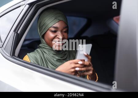 Sorridente Madonna musulmana nera utilizzando smartphone mentre si guida in auto Sedile posteriore Foto Stock