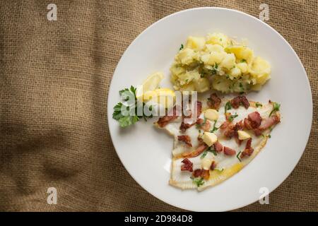 Un filetto di passera, Pleuronectes platessa, che è stato grigliato e servito con pezzi di pancetta alla griglia, prezzemolo, burro e patate bollite anche con p Foto Stock