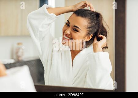 Bella donna sorridente che indossa accappatoio guardando lo specchio e regolando i suoi capelli Foto Stock
