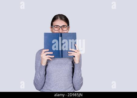 Felice studentessa con occhiali sorridenti coprendo mezza faccia con un libro blu aperto . Amante del libro. Pronto a studiare sodo. Foto Stock