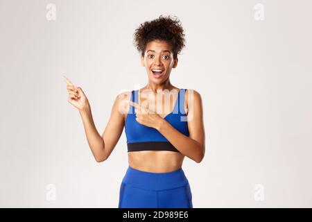 Sana e attraente sportivo in outfit blu sport, puntando le dita sinistra e guardando sorpreso, mostrando marchio o logo, sfondo bianco Foto Stock