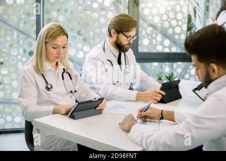 Team medico multietnico che ha una riunione in sala luce, lavorando su tablet digitali e prendere appunti. Focus sulla mezza età caucasica donna medico Foto Stock