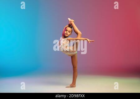 Energia. Piccola ragazza caucasica, allenamento di ginnastica ritmica, che si esibisce isolato su sfondo gradiente blu-rosso in studio al neon. Aggraziato e flessibile, bambino forte. Concetto di sport, movimento, azione. Foto Stock