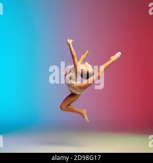 Salto alto. Piccola ragazza caucasica, allenamento di ginnastica ritmica, che si esibisce isolato su sfondo gradiente blu-rosso in studio al neon. Aggraziato e flessibile, bambino forte. Concetto di sport, movimento, azione. Foto Stock