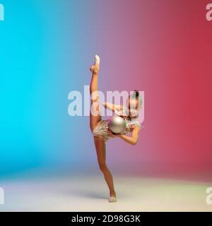 Energia. Piccola ragazza caucasica, allenamento di ginnastica ritmica, che si esibisce isolato su sfondo gradiente blu-rosso in studio al neon. Aggraziato e flessibile, bambino forte. Concetto di sport, movimento, azione. Foto Stock