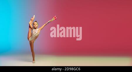 Volantino. Piccola ragazza caucasica, allenamento di ginnastica ritmica, che si esibisce isolato su sfondo gradiente blu-rosso in studio al neon. Aggraziato e flessibile, bambino forte. Concetto di sport, movimento, azione. Foto Stock