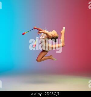 Salto alto. Piccola ragazza caucasica, allenamento di ginnastica ritmica, che si esibisce isolato su sfondo gradiente blu-rosso in studio al neon. Aggraziato e flessibile, bambino forte. Concetto di sport, movimento, azione. Foto Stock