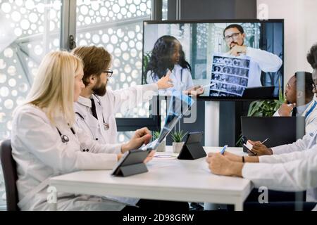 Team di medici multietnici che guardano lo schermo in videoconferenza incontro in ospedale, chiacchierando con due fiduciosi medici africani e caucasici Foto Stock