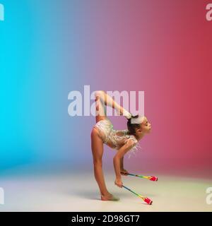 Offerta. Piccola ragazza caucasica, allenamento di ginnastica ritmica, che si esibisce isolato su sfondo gradiente blu-rosso in studio al neon. Aggraziato e flessibile, bambino forte. Concetto di sport, movimento, azione. Foto Stock