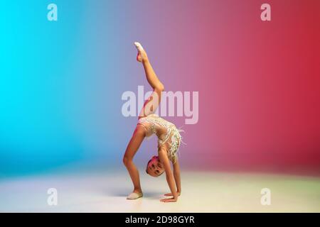 Dinamico. Piccola ragazza caucasica, allenamento di ginnastica ritmica, che si esibisce isolato su sfondo gradiente blu-rosso in studio al neon. Aggraziato e flessibile, bambino forte. Concetto di sport, movimento, azione. Foto Stock