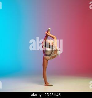 Dinamico. Piccola ragazza caucasica, allenamento di ginnastica ritmica, che si esibisce isolato su sfondo gradiente blu-rosso in studio al neon. Aggraziato e flessibile, bambino forte. Concetto di sport, movimento, azione. Foto Stock