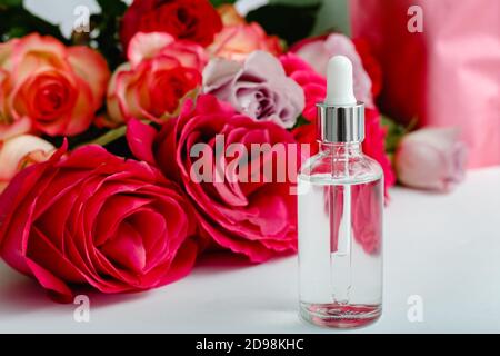 Siero su sfondo floreale tabella bianca. Fiore rosso rosa rose prodotto di bellezza naturale. Cura della pelle, trattamento del corpo. Bottiglia cosmetica in vetro con copia rosa Foto Stock
