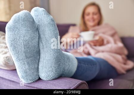 Donna a casa con calzini caldi e avvolti Coperta sdraiata su divano bere tazza di tè Foto Stock