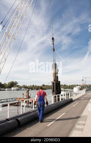 Belgrado, Serbia - 9 ottobre 2020: Gru di costruzione che solleva una grande punta industriale in acciaio per il pontile del fiume e un operaio, vista ad angolo basso riva del fiume Foto Stock
