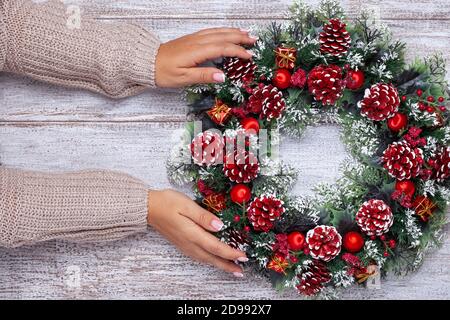 Due mani donna in maglia beige maglione con manicure francese fare wreath di Natale fatto in casa da coni, rami di albero, ornamenti su sfondo di legno Foto Stock