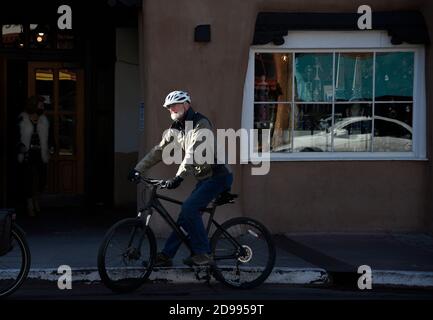 Un uomo anziano in visita a Santa Fe, New Mexico, in bicicletta si ferma lungo una strada ombreggiata del centro in un fascio di luce solare mattutina. Foto Stock