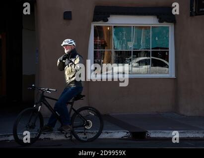 Un uomo anziano in visita a Santa Fe, New Mexico, in bicicletta si ferma lungo una strada ombreggiata del centro in un fascio di luce solare mattutina. Foto Stock