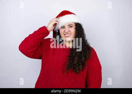 Giovane bella donna che indossa un cappello di Santa su sfondo bianco confondere e meraviglia circa domanda. Incerto con dubbio, pensando con mano sulla testa Foto Stock