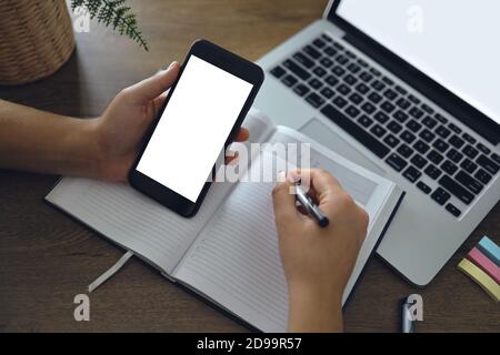 Immagine mockup della mano donna che tiene vuoto schermo bianco dello smartphone desktop con il computer portatile sul tavolo mentre scrive la sua idea sul notebook. Shopping online, Dig Foto Stock